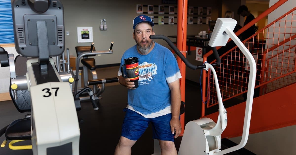 An individual with a blue t-shirt and blue shorts stands next to exercise equipment, holding a travel mug.