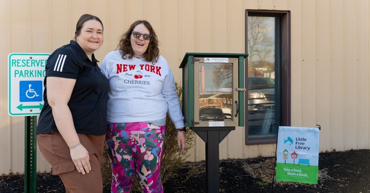 Two women stand and smile in front of a Little Free Library book box outside a building, next to a sign for accessible parking.