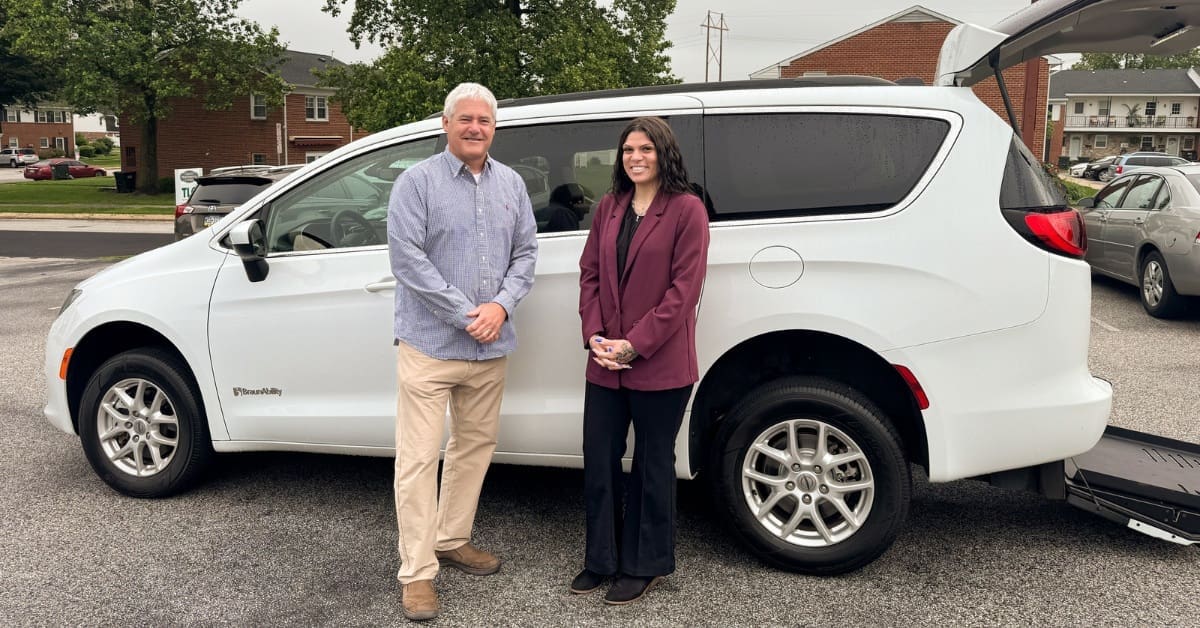 A man and a woman stand smiling in front of a white accessible minivan with a ramp, parked in a lot.