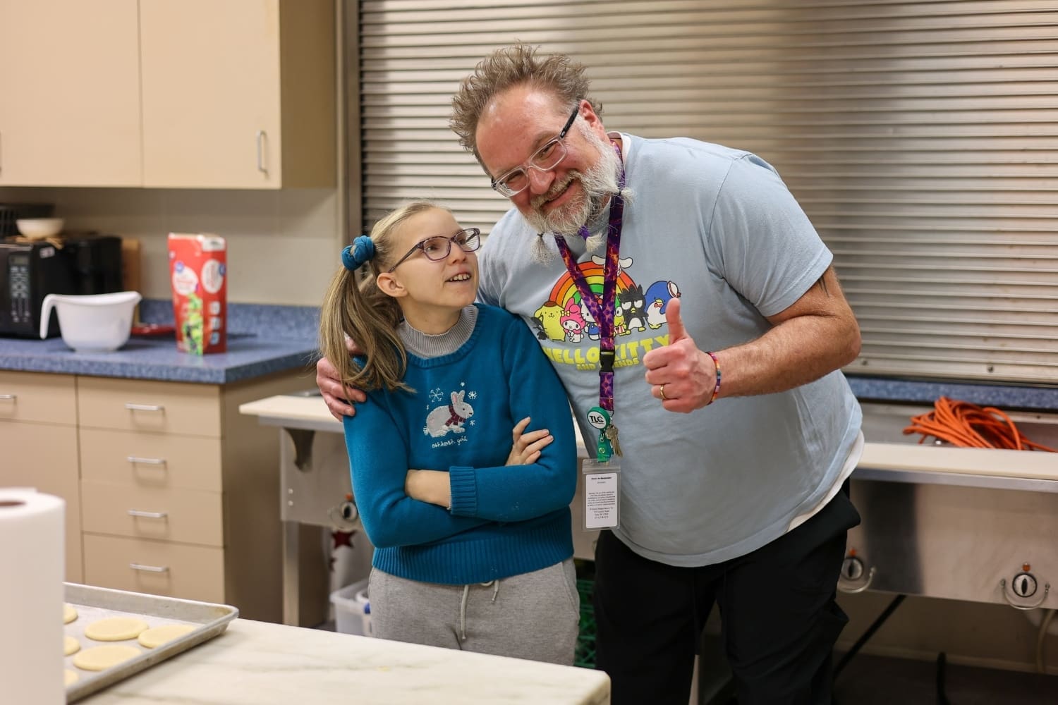 A TLC staff member stands beside a program participant in a kitchen space, smiling and giving a thumbs-up during a baking activity.