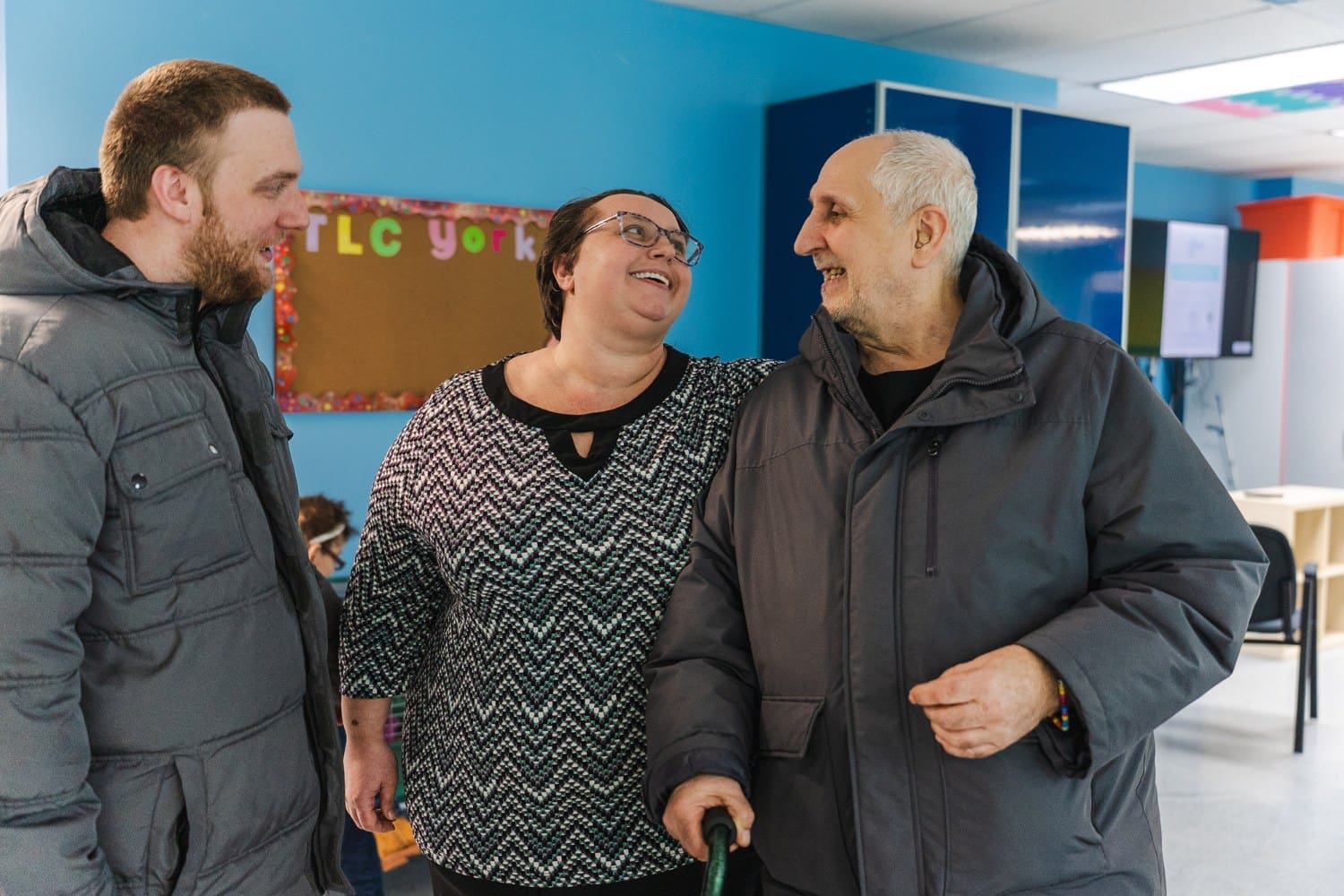 Program Specialist Amanda Burg stands between two men inside a TLC facility, smiling and talking with them in a welcoming, supportive setting.